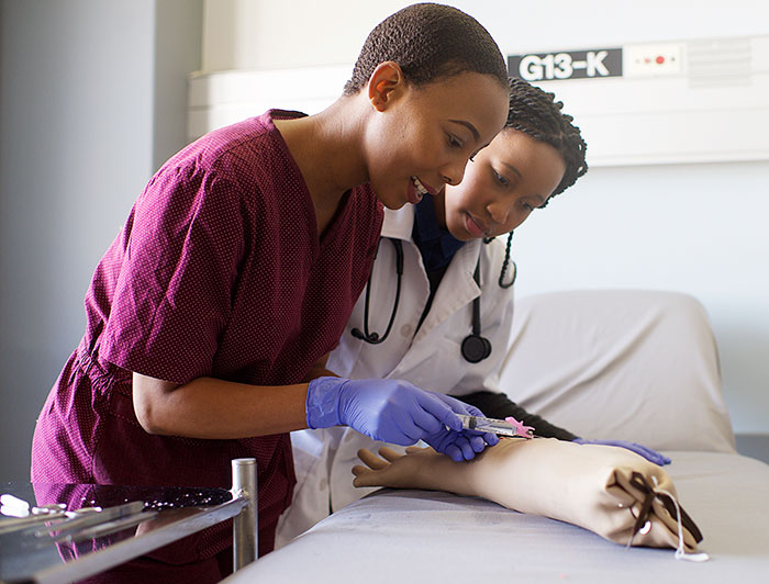 UMass Memorial Health. A black female clinician demonstrates injection on a teaching prosthetic while apprentice looks on