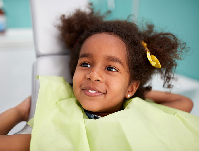 Barbara Bush Children’s Hospital/MaineHealth. A young girl with afro puffs and yellow bows sits smiling in a dentist chair