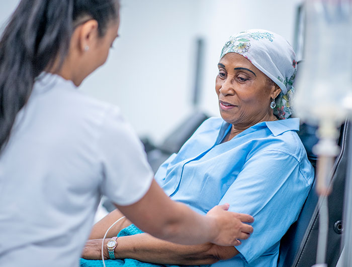 The Queen’s Health Systems. An older woman wearing a scarf on her head sits in a chair receiving cancer treatment from a nurse