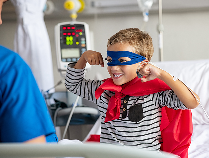 Unity Point - Blank Children's Hospital. Stock image: A young patient sits in a hospital bed flexing his superhero muscles, wearing a red superhero cape and blue mask