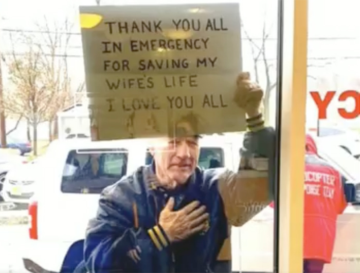 Telling the Hospital Story: Gratitude for Lifesaving Care at Morristown Medical Center; A man stands outside an emergency room window holds a sign reading: Thank you all in Emergency for saving my wife's life. I love you all.