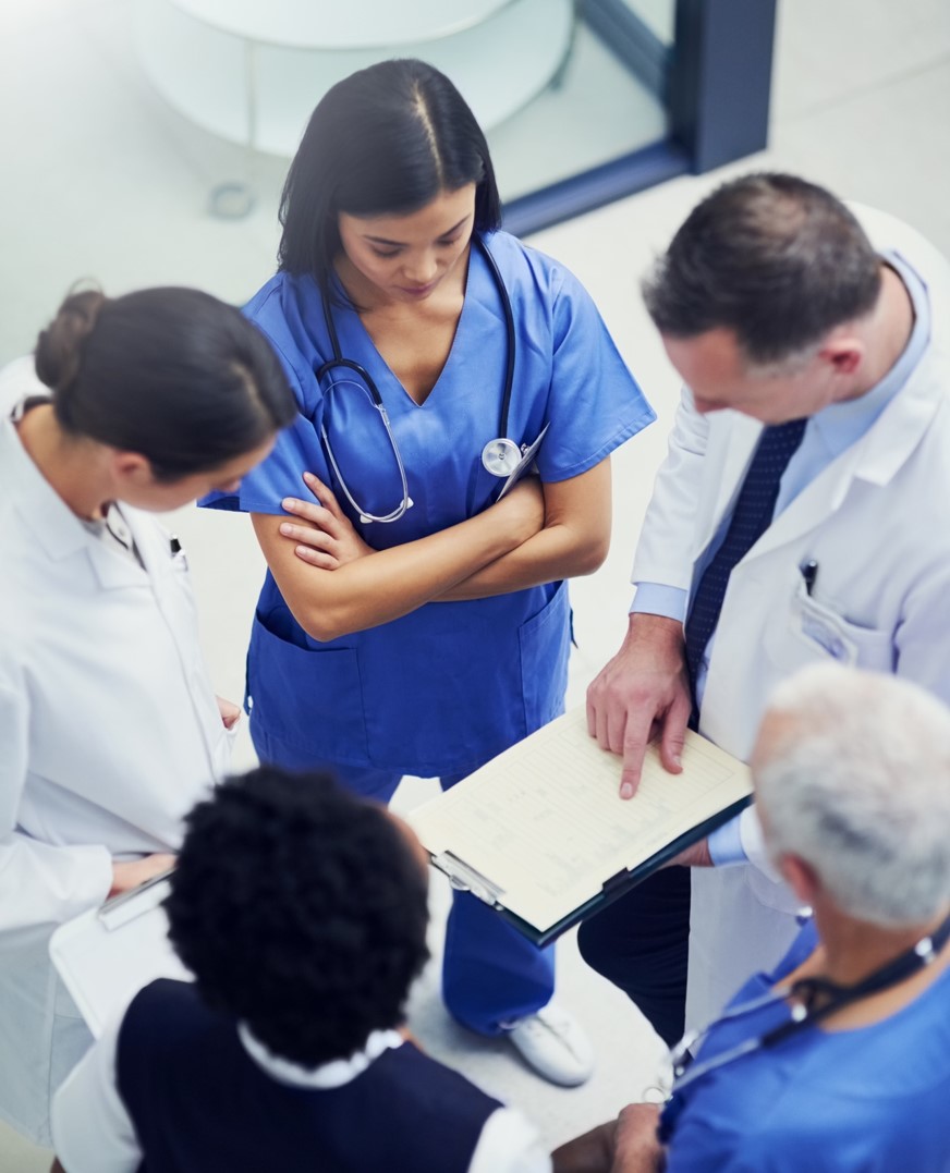 Clinicians gathered in a circle reviewing information on a clipboard held by a physician.