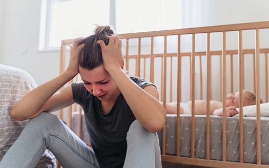 Mayo Clinic to Study Digitized Workflows, AI-Backed Messaging to Improve Postpartum Care. A mother holds her head in her hands while sitting on the floor while her baby sleeps in a crib behind her.