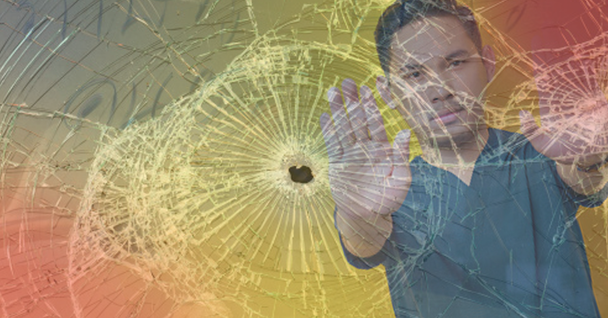 Navigating the Rising Tide of Violence in Health Care Environments. A clinician in scrubs stands with his arms extended and palms up behind a shattered window with a bullet hole in it.