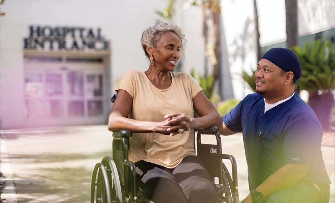 A patient in a wheelchair accompanied by a clinician in scrubs enjoys the sunshine outside of a ScionHealth hospital entrance.