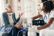 Amazon, Intermountain and Ascension Lead Push to Expand Home-Based Care. A seated home-health nurse wearing a surgical mask and nitrile gloves speaks with a patient wearing an oxygen mask in the patient's home.