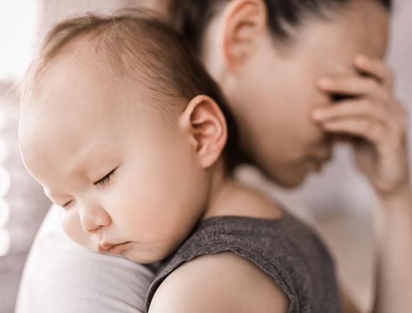 Stock photo of a mother holding her forehead with one hand, supporting a sleeping infant on her shoulder with the other