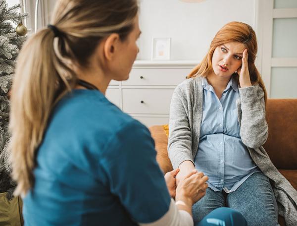 A pregnant woman sits on a sofa talking with a female clinician