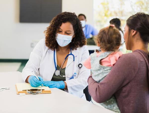 A female physician wearing medical mask sits filling out forms with a mother and child