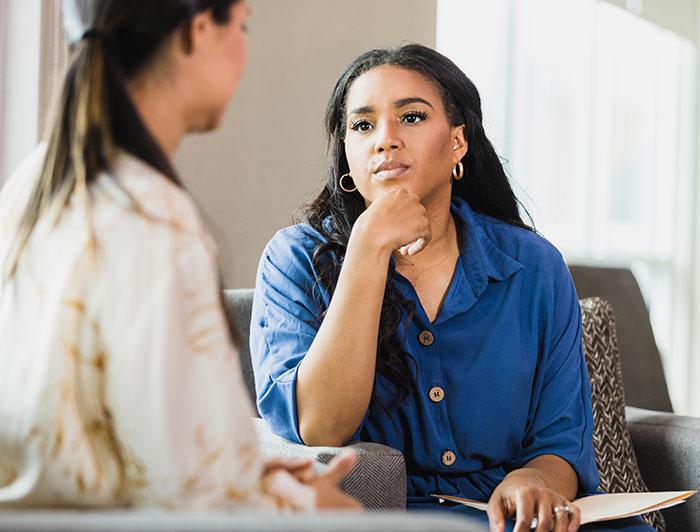Two people sitting down in an office. One is holding a folder with notes, and is listening to the other person. 
