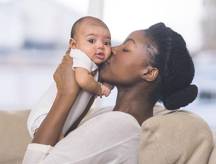 An african american mother, eyes closed, kisses her newborn baby on the cheek. Baby is looking towards viewer.