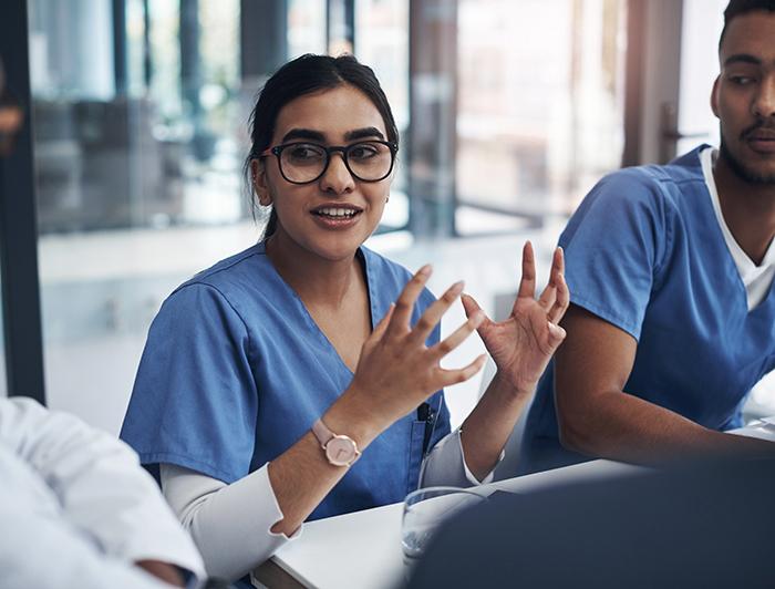 Woman wearing scrubs and glasses talks and gestures with her hands as she sits in a group of peers 