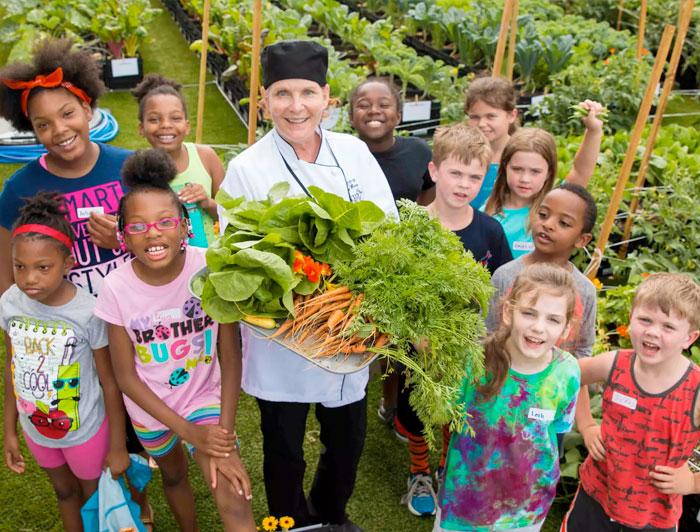 A BMS chef stands in a garden holding produce, surrounded by smiling neighborhood children.