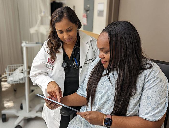A femal Loma Linda University Medical Center clinician in white coat stands reviewing a tablet with a fmale patient wearing hospital gown