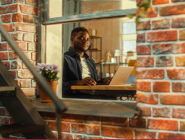 A young black sitting in an apartment at a table working on a laptop, viewed through a fire escape window