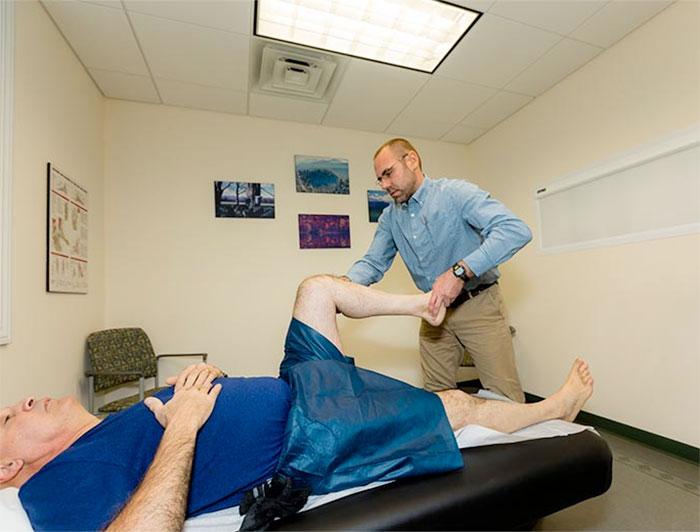A clinician manipulates the leg of an elderly man lying prone on an examination table