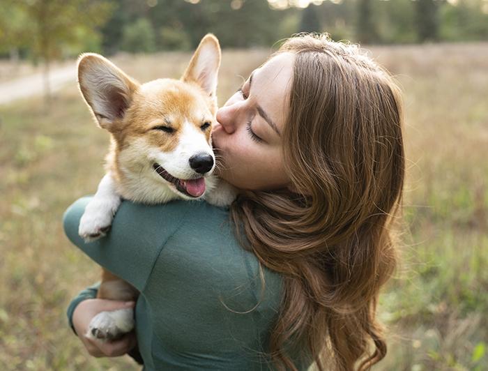 A young woman stands in a field holding and kissing a Corgi puppy