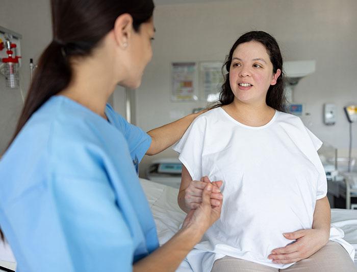 A pregnant woman sits on the edge of a hospital bed holding the hand of a woman in scrubs