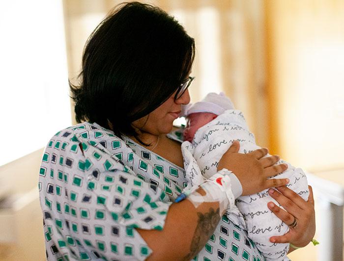 Mother in hospital gown sits cradling swaddled newborn to her chest