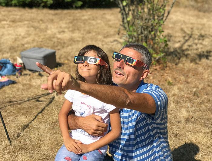 A man and child wearing solar eclipse glasses look up at the eclipse