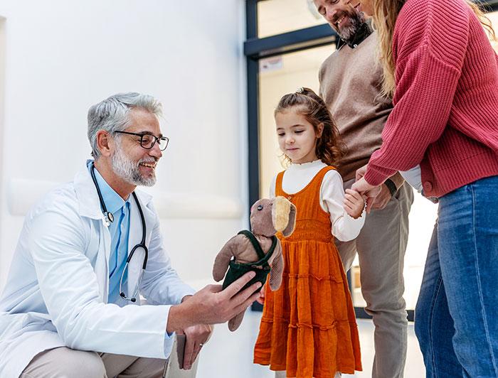 A male clinician crouches next to a little girl and her parents, offering the child a teddy bear