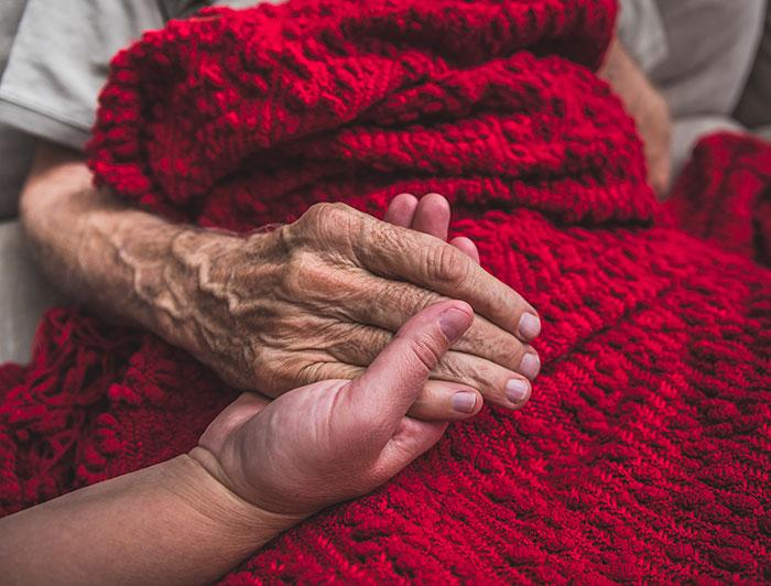 a younger person holds holding the hand of an elderly person sitting under a knitted blanket