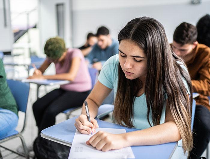 A young woman sits in a classroom at a desk taking a test,  among a class of students doing the same