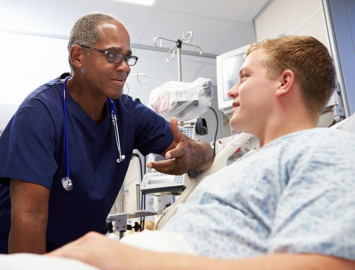 A male clinician sits bedside, talking to a male patient in hospital bed