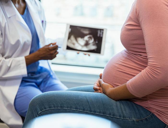 Stock. A female physician in scrubs and white coat sits showing ultrasound photo to an expecting mother.