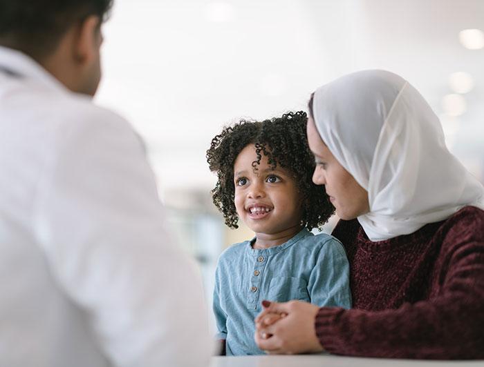 A smiling, curly haired child and mother in head scarf sit talking to a white coated physician 