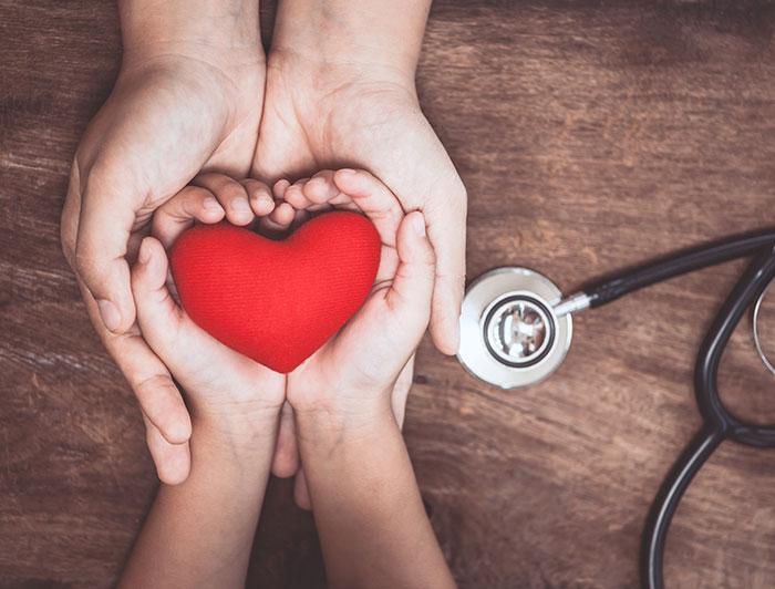 stock image of adult hands holding a child's hands holding a red heart. A stethoscope sits on the table.