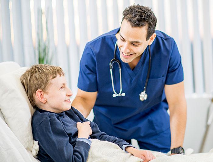 A male physician in scrubs smiles at a frail boy sitting up in a hospital bed