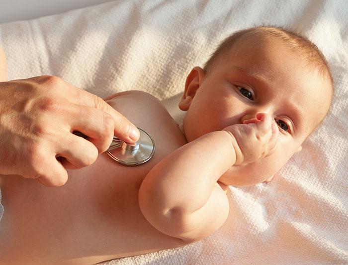 An infant rests on an exam table while a hand places a stethoscope against the infants chest.