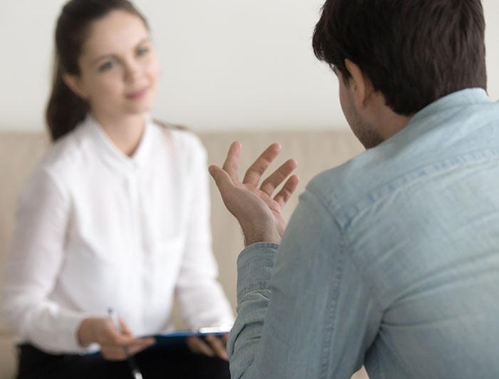 A female counselor holding a clibpoard and pen sits listening to a male patient