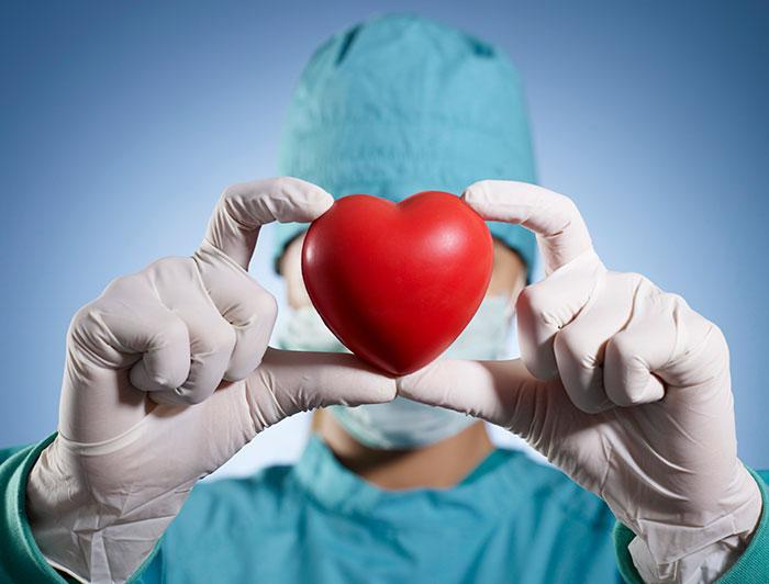 Stock image shows a surgeon in scrubs holding a toy heart aloft, masking face
