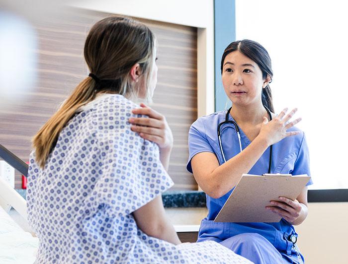 A female nurse in scrubs talks with a female patient sitting in a hospital bed