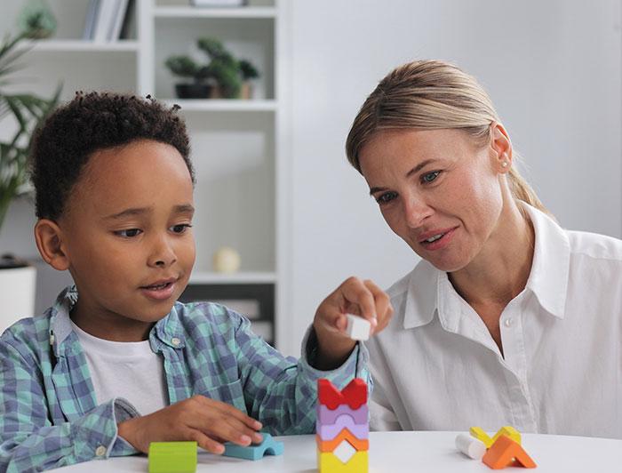 A young boy sits stacking blocks as a carer looks on