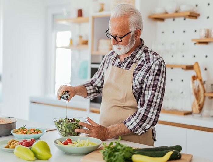 An older man wearing an apron stands in a kitchen preparing health food