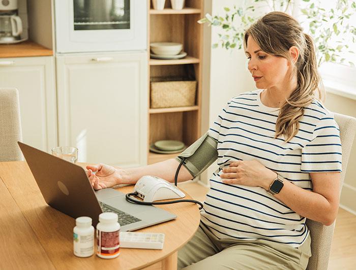 A pregnant woman sits at a kitchen table measuring her blood pressure and 