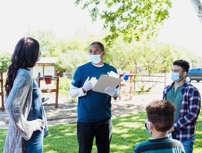 A male healthworker wearing PPE and holding a clipboard stands outdoors talking with a woman, a man and a child