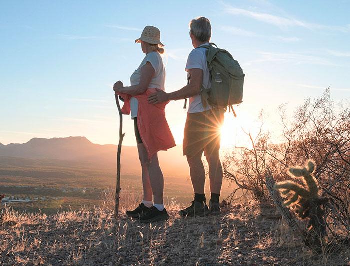 A couple in hiking gear stands on a trail, looking towards a sunrise