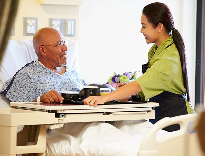 An older man in a hospital bed exchanges a smile with a hospital worker serving a hospital meal