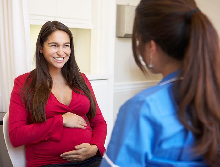 A smiling pregnant woman sits cradling her belly while talking with a female nurse