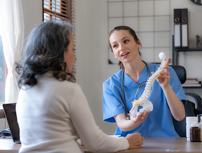 A  female doctor in scrubs holds a model spine while talking with a woman